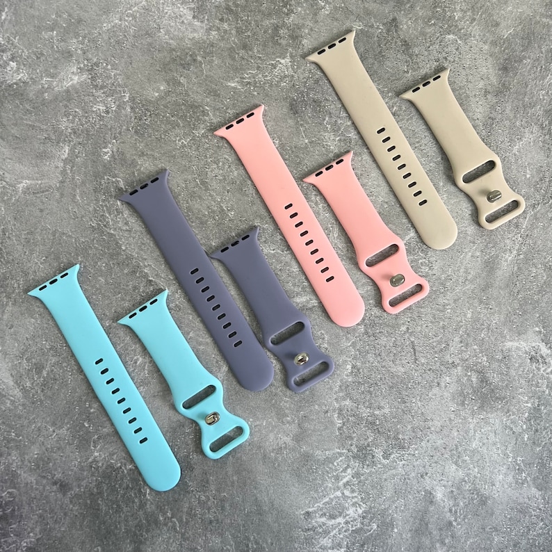 May include: A set of six silicone watch bands in various colors, including light blue, pink, gray, and purple. The bands are designed for Apple Watch and have a buckle closure.