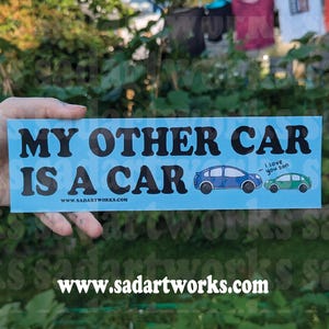 以下が含まれることがあります： 「MY OTHER CAR IS A CAR」の黒い文字が入った水色のバンパーステッカー。青と緑の車のイラストと「I love you son」の文字が特徴です。ウェブサイトwww.sadartworks.comもステッカーに印刷されています。