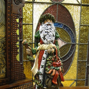 May include: A Santa Claus figurine wearing a green and red coat with white fur trim, a matching hat, and a white beard. The figure holds a wooden staff in one hand and a woven basket filled with greenery, red berries, and carrots in the other.