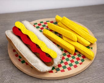 May include: A felt hot dog with red and yellow toppings, and a side of yellow felt fries, presented on a wooden plate with a red and green checkered pattern. The hot dog is in a white bun.