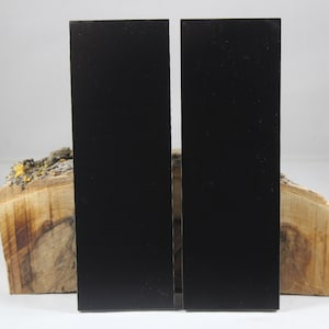 May include: Two black rectangular knife scales, made of a smooth material, are displayed on a piece of wood with a natural bark texture.