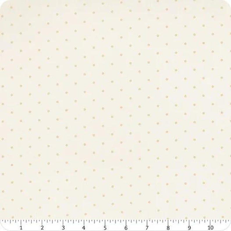 May include: A piece of off-white fabric featuring a repeating pattern of small, light tan polka dots. A ruler is visible along the bottom edge, showing a measurement scale from 0 to 10 inches, indicating the width of the textile material.