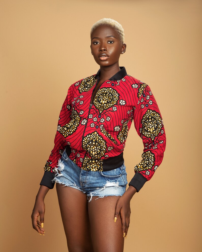 Puede incluir: Una chaqueta bomber de color rojo y dorado con cremallera negra y ribete negro. La chaqueta se lleva sobre unos pantalones cortos de mezclilla.