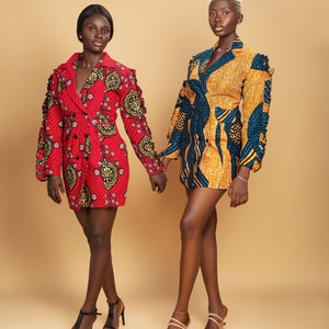 May include: Two women wearing short, patterned dresses with long sleeves and collars. The dresses have a blazer style and are made of a colorful fabric. The women are standing side by side and holding hands. They are both wearing high heels.