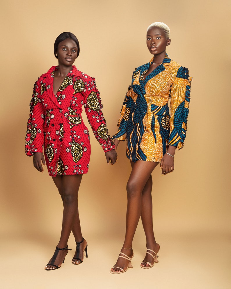 May include: Two women wearing short, patterned dresses with long sleeves and collars. The dresses have a blazer style and are made of a colorful fabric. The women are standing side by side and holding hands. They are both wearing high heels.
