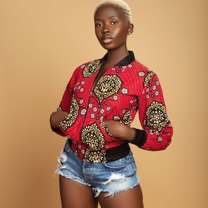 Puede incluir: Una mujer con el pelo corto rubio lleva una chaqueta bomber roja y dorada con estampado, con una cremallera negra y ribetes negros. Tambi&eacute;n lleva pantalones cortos de mezclilla con detalles desgastados.