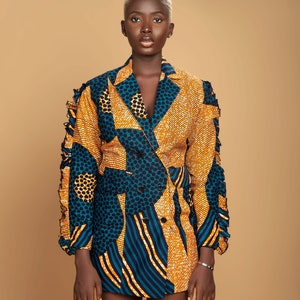 May include: A woman wearing a short-sleeved blazer with a geometric pattern in shades of blue, yellow, and black. The blazer has a double-breasted closure and a ruffled trim on the sleeves.