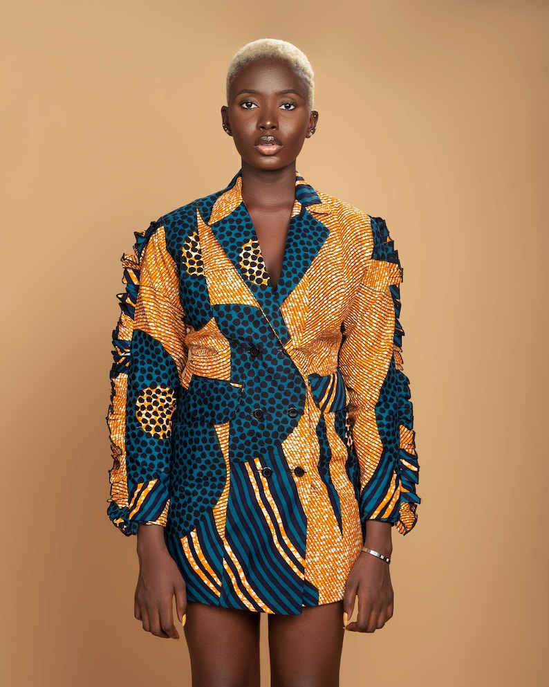 May include: A woman wearing a short-sleeved blazer with a geometric pattern in shades of blue, yellow, and black. The blazer has a double-breasted closure and a ruffled trim on the sleeves.