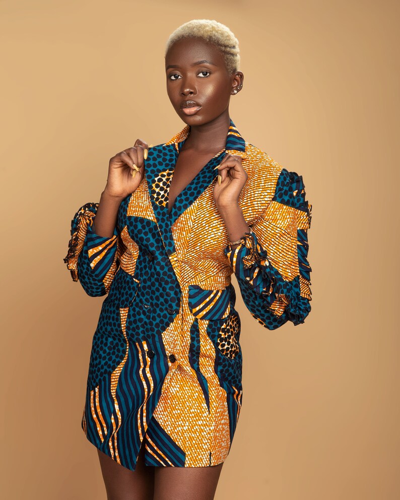 May include: A woman wearing a short, double-breasted blazer with a colorful geometric pattern in shades of blue, yellow, and orange. The blazer has a ruffled detail on the sleeves.