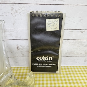 May include: A black Cokin Filter/Exposure Record booklet with the text "FILTER/EXPOSURE RECORD and Model Releases". The booklet is in a clear plastic sleeve. A handwritten sticker reads "0000 1719 Chris Ward" and "$ 2.49".