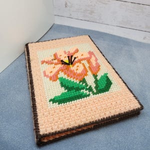 May include: A pink and green embroidered flower design on a pink fabric background. The fabric is framed with a brown border.