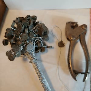 May include: A bundle of silver security tags with a silver tag applicator tool. The tags are attached to a string and have a small, round, silver disc at the end.