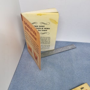 May include: An open vintage book titled "The Diet for People Who Want to Diet" with a "Carbohydrate Gram Counter" on the cover. The book is orange and cream-colored, with text and illustrations. A metal ruler is next to the book.
