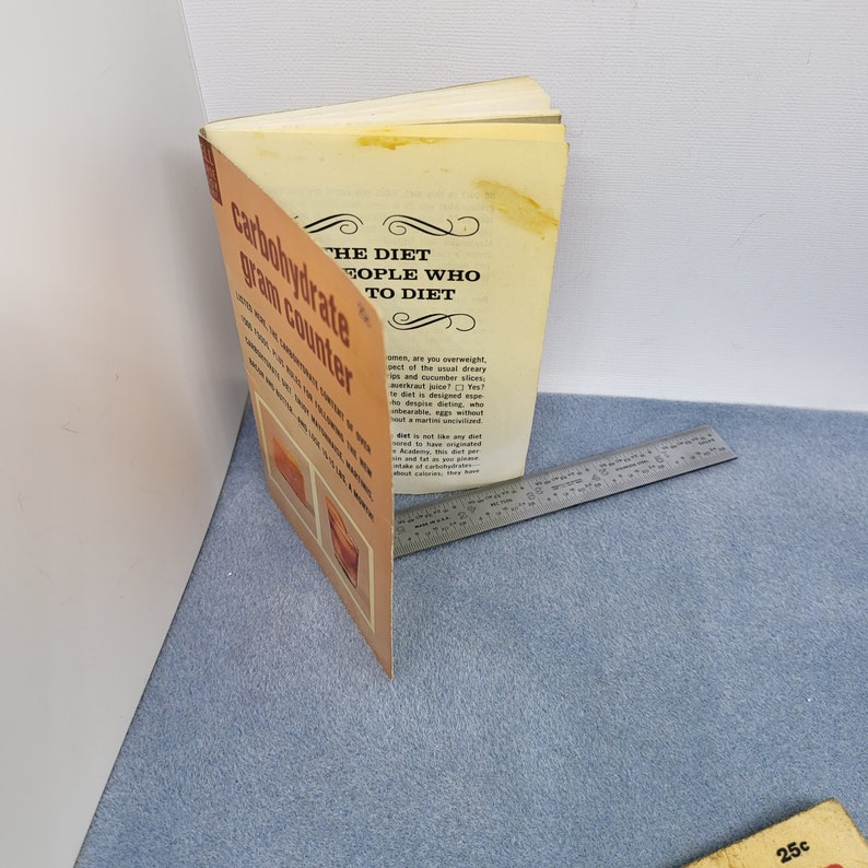 May include: An open vintage book titled "The Diet for People Who Want to Diet" with a "Carbohydrate Gram Counter" on the cover. The book is orange and cream-colored, with text and illustrations. A metal ruler is next to the book.