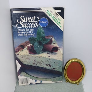 May include: A vintage Pillsbury "Sweet Success" cookbook with a dessert image on the cover, and a small gold-colored oval picture frame. The cookbook has a dark blue cover with white text and the Pillsbury logo.