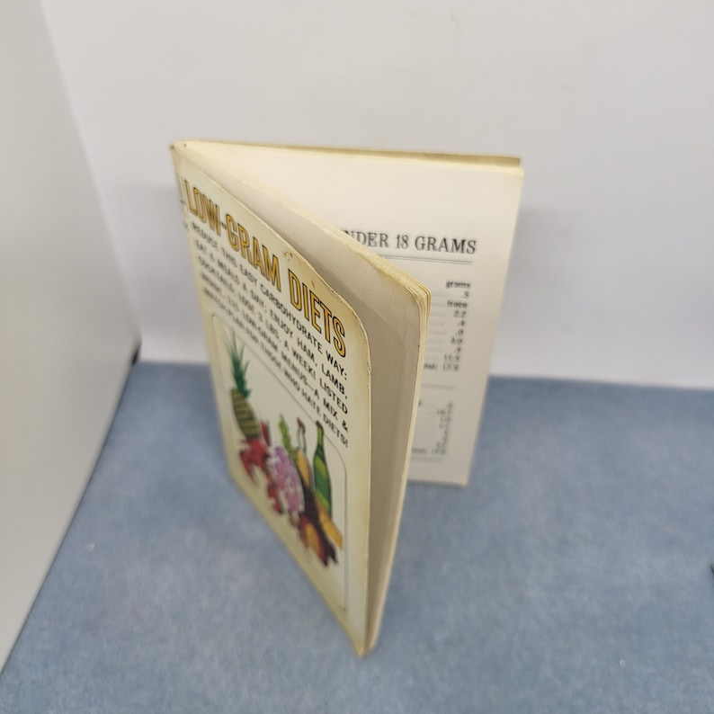 May include: An open book titled "Low-Gram Diets" with text and illustrations of fruits and vegetables. The book's cover features a pineapple, flowers, and bottles. The pages are yellowed with age, and the book is set against a light background.