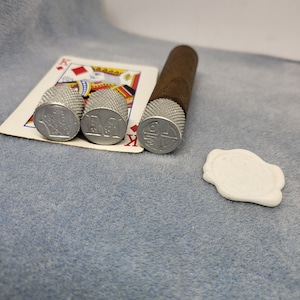 May include: A brown cigar with a silver cap rests next to three silver thimbles with embossed designs and a playing card. A white wax seal is also present. The items are on a blue surface.