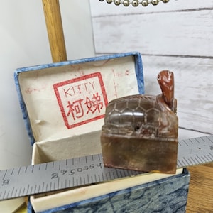 May include: A brown turtle-shaped stamp with a red head and a rectangular base, next to an open blue box with the word "KITTY" and Chinese characters in red. A metal ruler is also present.