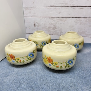 May include: Four small, cream-colored ceramic jars with floral designs. The jars have a raised rim and are decorated with a band of orange, yellow, blue, and white flowers.