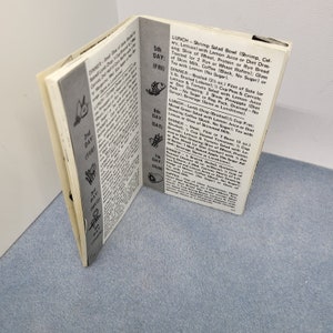 May include: An open booklet with printed text and illustrations, likely a diet or meal plan guide. The pages are off-white with black text and graphics, and the booklet is slightly angled, showing the interior pages.