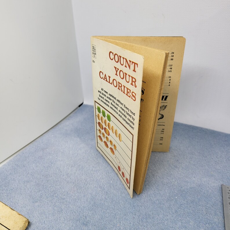 May include: A vintage paperback book titled "Count Your Calories" with a beige cover and brown text. The cover features a graphic of food items and a list of calories. The book is open, revealing interior pages with calorie information.