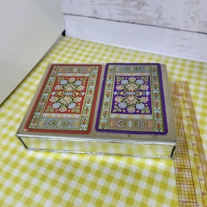 May include: A vintage playing card set in a silver-colored metal box. The cards feature ornate designs in red, purple, and gold. The box is rectangular and sits on a yellow and white checkered surface.