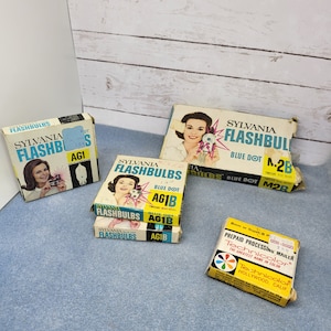 May include: Vintage Sylvania Flashbulbs boxes with retro graphics and a Technicolor prepaid processing mailer. The boxes feature images of a woman with a camera and the text "FLASHBULBS". The mailer is yellow with a colorful Technicolor logo.