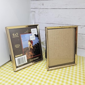 May include: Two gold-colored metal photo frames. One frame displays a photo of a couple on a mountain, labeled "Horizons." The other frame is empty. Both frames are 4x5 inches and can be displayed vertically or horizontally.
