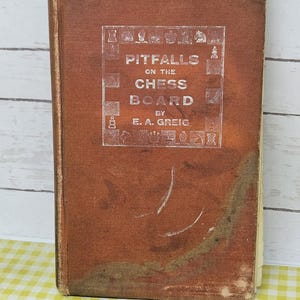 May include: A vintage book titled "Pitfalls on the Chess Board" by E.A. Greig. The cover is a worn, reddish-brown color with the title and author's name in white lettering. Chess pieces are illustrated around the title.