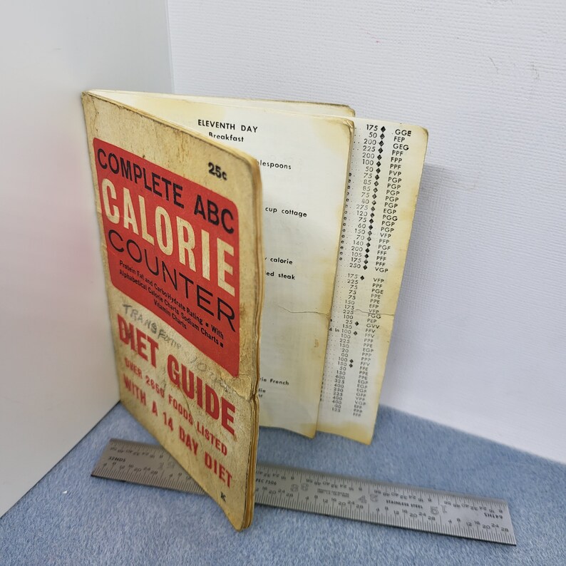 May include: Vintage "Complete ABC Calorie Counter Diet Guide" book. The cover is red and cream-colored with the title in bold letters. The book is open to a page with a list of foods and calorie counts. A metal ruler is next to the book.