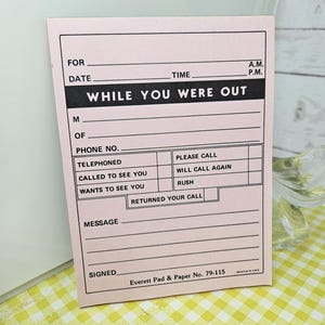 May include: A pink "While You Were Out" message pad with black text. The pad includes fields for date, time, and contact information. The text "Everett Pad & Paper No. 79-115" is printed at the bottom. The pad is on a yellow and white checkered surface.
