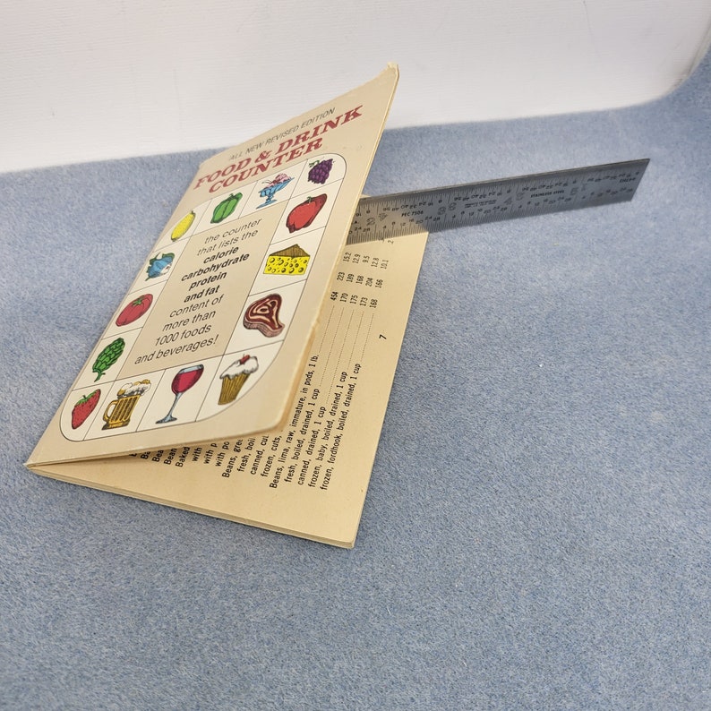 May include: An open book titled "FOOD & DRINK COUNTER" with illustrations of various foods and beverages. The book lists the calorie, carbohydrate, protein, and fat content of over 1000 foods. A metal ruler is placed across the pages.