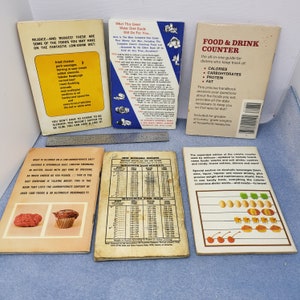May include: A collection of vintage diet books and guides, including a "Food & Drink Counter" and a "Water Diet Guide." The books feature lists of foods and calorie information, with a metal ruler in the background.