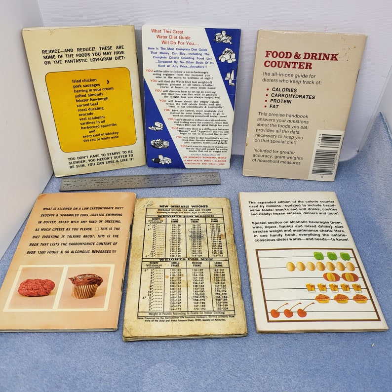 May include: A collection of vintage diet books and guides, including a "Food & Drink Counter" and a "Water Diet Guide." The books feature lists of foods and calorie information, with a metal ruler in the background.