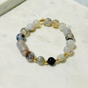 Könnte beinhalten: Ein Armband aus weißen Achatperlen mit goldenen Akzenten und einer schwarzen Onyxperle. Das Armband liegt auf einer weißen Oberfläche.