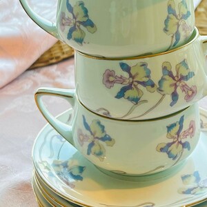 May include: A stack of light green teacups and saucers with a floral design in blue, yellow, and purple. The cups have gold trim and handles. The saucers are stacked below the cups.