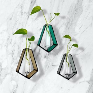 May include: Three geometric wall vases with glass tubes holding green plants. The vases have diamond-shaped wooden frames in shades of brown, green, and gray. The background is a white marble wall.