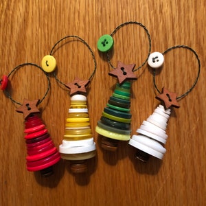 May include: Four handmade Christmas tree ornaments made from buttons. The trees are decorated with red, yellow, green, and white buttons. Each tree has a wooden star on top and is attached to a string.