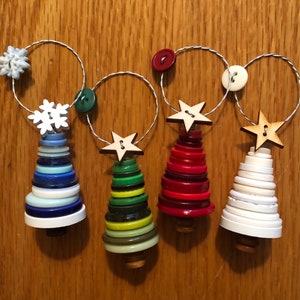 May include: Four handmade Christmas tree ornaments made from stacked buttons. The trees are decorated with wooden stars and are strung together with wire. The trees are blue, green, red, and white.