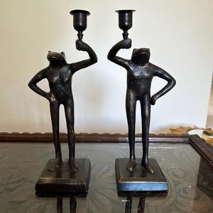 May include: Two bronze frog-shaped candle holders. Each frog figure stands on a rectangular base, holding a candle cup above its head. The figures have a textured finish and are positioned on a reflective surface.
