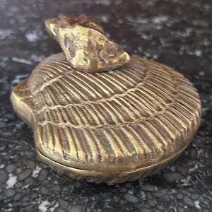 May include: A vintage brass seashell-shaped trinket box with a hinged lid. The box has a detailed, textured surface and a small, irregular-shaped handle on top. The box is displayed on a dark, marbled surface.