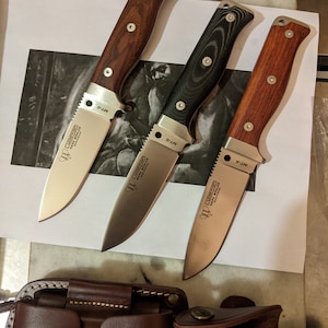 May include: Three hunting knives with wooden handles in shades of brown and black, and two brown leather sheaths. The silver blades have the text "Cudeman NESS BOWLER" and "MT-5". These knives are designed for outdoor activities and general use.