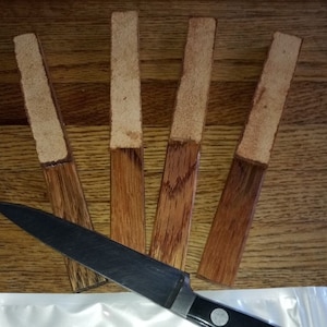 May include: Five wooden strips with a light brown finish, each with a section of light brown leather attached. A black kitchen knife is placed in front of the strips.