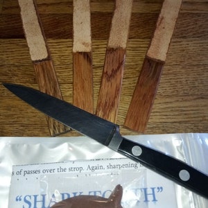 May include: Five wooden sharpening sticks with leather strips on top. The sticks are arranged in a fan shape. A knife and a small chocolate fish are in the foreground. The text "SHARPATH" is visible on a white background.