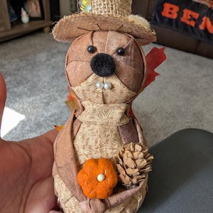 May include: A fabric beaver ornament, wearing a straw hat with a bee, holding a small orange pumpkin and a pinecone. The beaver has black eyes, a black nose, and two white pearl teeth. The figurine is made of various brown and tan fabrics.