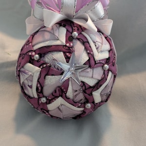 Pode incluir: Um enfeite de Natal artesanal com um adorno em forma de estrela. O enfeite é uma esfera, coberta com um patchwork de tecido roxo e branco. É adornado com pérolas falsas e encimado por um laço de fita lavanda e prateada.