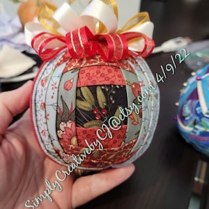 Pode incluir: Um ornamento esférico de tecido com um design de patchwork em tons de vermelho, verde e creme com motivos florais e de azevinho. O ornamento é rematado com um laço decorativo de fitas sobrepostas em branco, dourado e vermelho. O texto na imagem diz Simply CreativebyCJ@etsy.com 4/9/22.