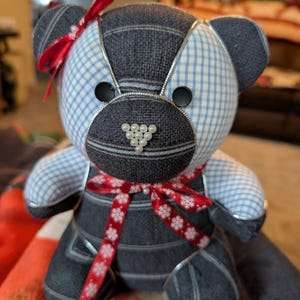 May include: A handmade teddy bear with a patchwork design. The bear features a mix of blue and gray fabrics, including a gingham pattern and denim-like material. It has black button eyes, a pearl bead nose, and is adorned with red ribbons.