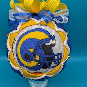 Könnte beinhalten: Ein blauer und gelber Stoffschmuck mit einem Rams-Logo und einer gelben Schleife oben. Der Schmuck steht auf einem klaren Glasständer.