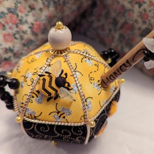 May include: A yellow and black bee pincushion with a gold trim and a white pearl top. The pincushion is shaped like a beehive and has a small bee on the front. The pincushion is decorated with a wooden stick with the word "Honey" written on it.